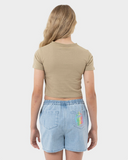 A young woman viewed from the back wearing a beige short-sleeve crop top and light blue denim shorts with a colorful graphic on the pocket, set against a plain light gray background.